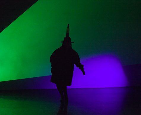 Auf einer Bühne ist der Schatten einer Figur zu sehen, die vor grüner und blauer Beleuchtung einen Hut mit einer Flasche auf dem Kopf trägt.